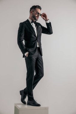 attractive elegant man in tuxedo with hand in pocket arranging sunglasses and looking to side on grey background