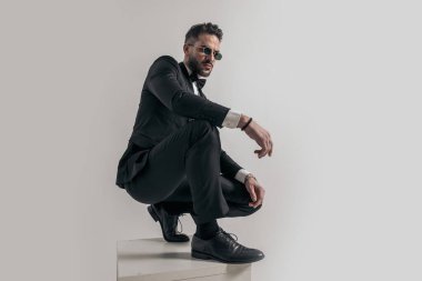 side view of elegant groom with sunglasses crouching on top of wooden box on grey background in studio