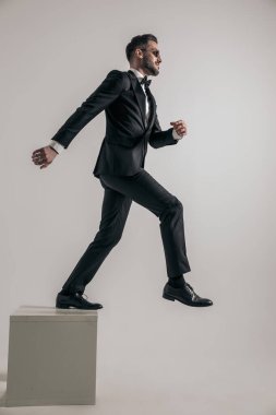 side view of elegant man in tuxedo stepping out of wooden box and starting a new life on grey background in studio