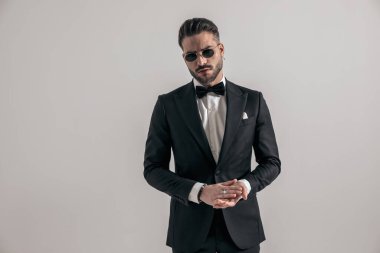 handsome businessman wearing elegant tuxedo and rubbing palms while posing in front of grey background in studio