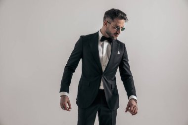 portrait of sexy elegant groom wearing black tuxedo and looking to side while posing in front of grey background in studio