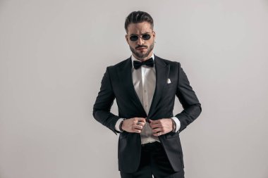 elegant young groom with sunglasses unbuttoning tuxedo and posing in front of grey background in studio