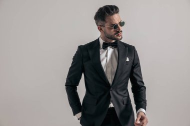 attractive young man wearing elegant tuxedo looking to side and holding hand in pocket on grey background in studio