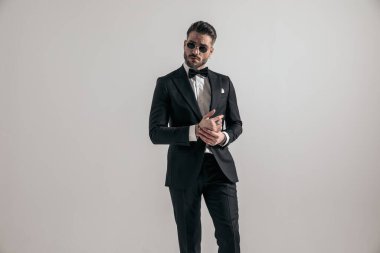elegant businessman wearing black tuxedo and touching palms while posing in front of grey background