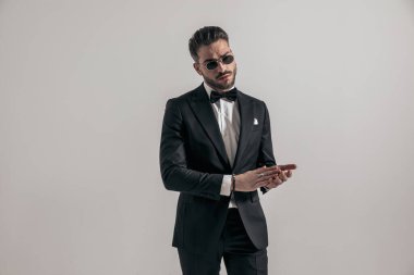 portrait of handsome unshaved guy wearing elegant tuxedo rubbing and touching palms on grey background in studio