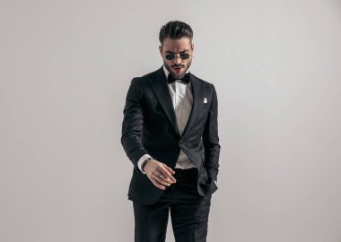 sexy young groom with glasses looking dowwn and holding hand in pocket on grey background in studio