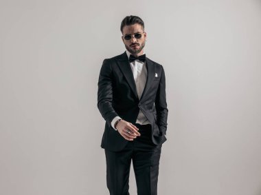 handsome elegant man with hand in pocket wearing black tuxedo and posing on grey background