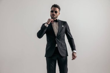 irresistible man in black tuxedo fixing bowtie while confidently posing in front of grey background in studio
