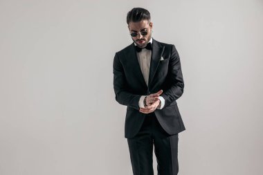 cool elegant man in black tuxedo looking down and rubbing palms in front of grey background in studio