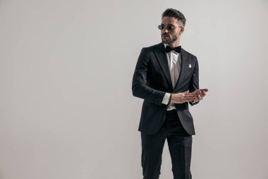 sexy elegant groom with sunglasses wearing black tuxedo and touching palms while posing in front of grey background in studio