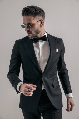 attractive young man in black tuxedo posing in a fashion way while looking to side in front of grey background in studio