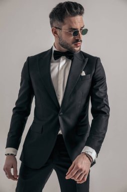 elegant groom with sunglasses in black tuxedo looking to side and posing in front of grey background