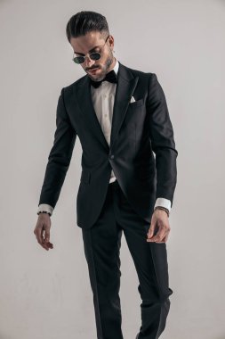 sexy unshaved guy in black tuxedo looking down and posing in front of grey background
