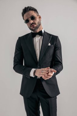 attractive elegant man rubbing palms and wearing black tuxedo, confidently posing in front of grey background in studio