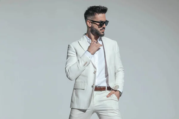 bearded man adjusting open collar shirt, wearing white suit for white party and looking to side, posing with hand in pocket in studio