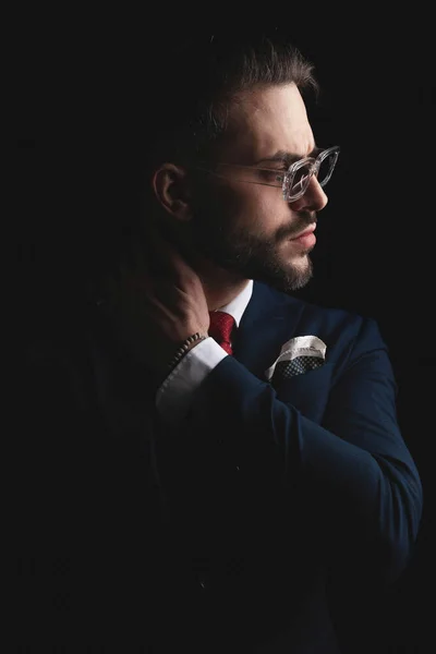 bearded elegant guy in suit with sunglasses holding hand behind neck and looking to side while posing in a cool and mysterious manner on black background in studio