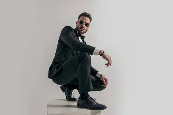 crouched businessman in tuxedo holding elbow on knee and posing in front of grey background on top of wooden box