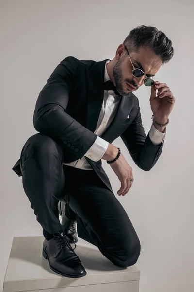 crouched businessman in elegant tuxedo arranging sunglasses, looking down, holding elbow on knee and posing on grey background in studio
