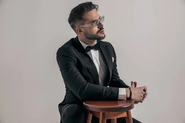 crouched sexy young man wearing tuxedo and sunglasses looking away while holding arm on wooden chair on grey background in studio