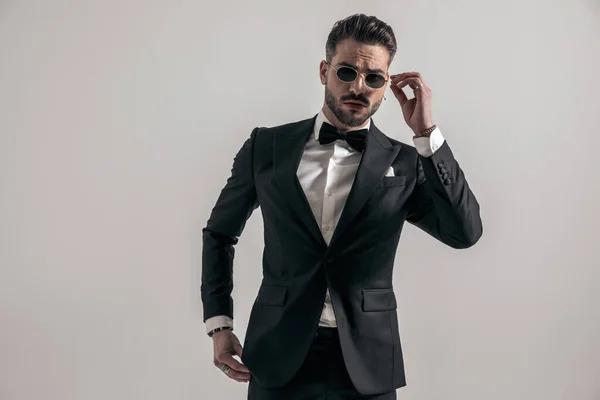 portrait of attractive bearded man in elegant tuxedo fixing sunglasses and posing in front of grey background