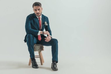 Şık takım elbiseli seksi bir adam dizinin üstünde dirseğini tutuyor ve stüdyoda gri arka planda kürk sandalyesine oturuyor.