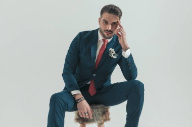 Takım elbiseli düşünceli genç adam el ele tutuşuyor ve gri arka planda kürk sandalyesinde oturuyor.