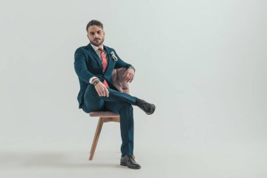 Sakallı, takım elbiseli, bacağını dizinde tutan ve stüdyoda gri arka planda poz veren kendine güvenen şık bir adam.