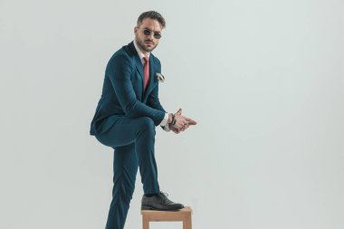 Zarif takım elbiseli, kendine güvensiz bir adamın portresi. El ele tutuşmuş, dirseğini dizinde, bacağını sandalyede tutuyor ve poz veriyor. 