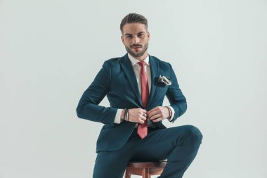 Sakallı mutlu genç iş adamı lacivert takımını ilikliyor, tahta koltukta oturuyor ve gri arka planda kendine güvenen bir şekilde poz veriyor.