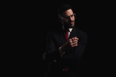 Gözlüklü, kollarını kavuşturup stüdyoda esrarengiz bir şekilde siyah arka planda poz veren takım elbiseli zarif bir adam.