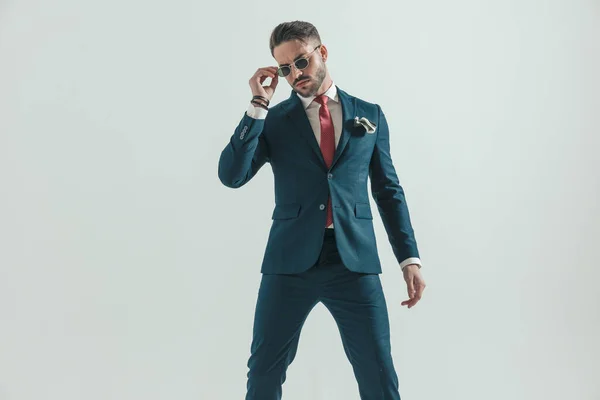 portrait of cool fashion man in elegant suit posing in a confident way while fixing sunglasses in front of grey background in studio