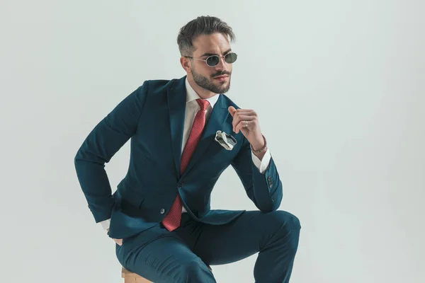 cool young businessman with beard wearing retro sunglasses and holding elbow on knee, smiling and looking away in studio