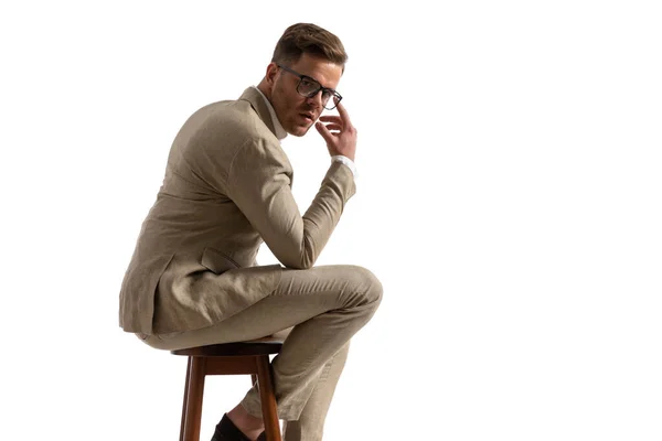 在演播室里 一位有魅力的商人坐在木椅上 在白色背景前摆姿势 调整太阳镜的侧视图 — 图库照片