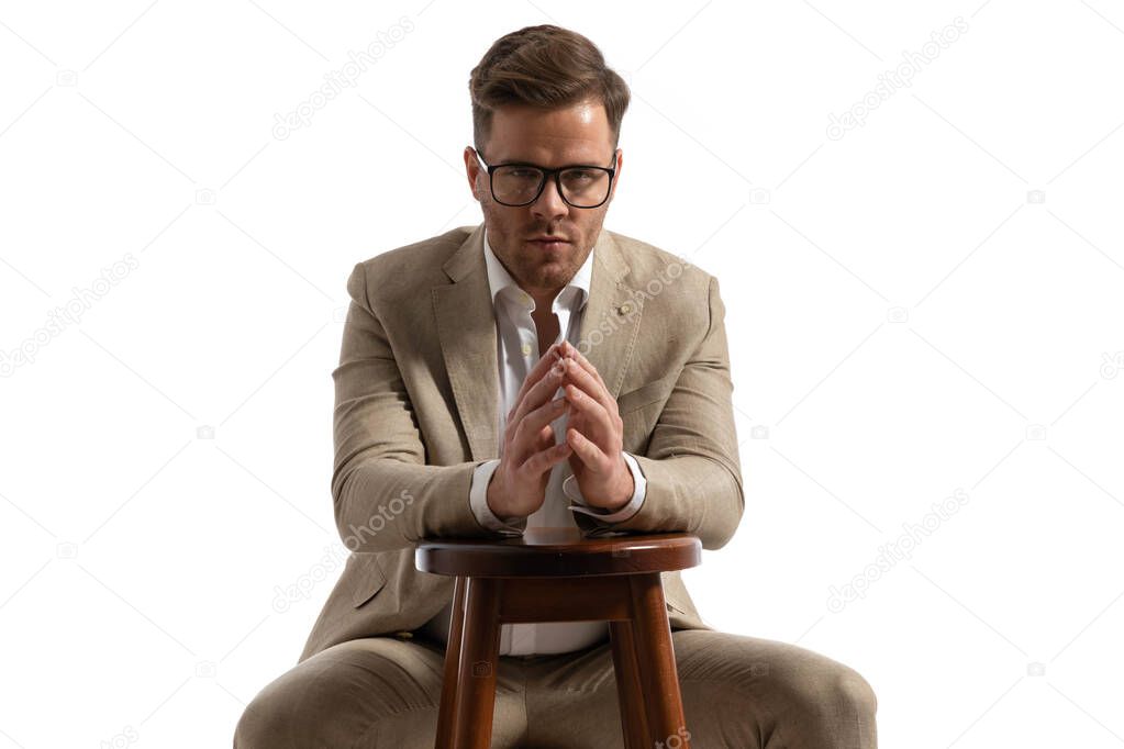 young man with glasses holding hands together on wooden chair and posing in front of white background in studio