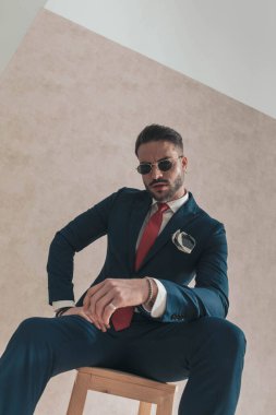 Şık takım elbiseli seksi adam dirseğini dizinde tutuyor ve aşağıdan kendine güvenen bir şekilde poz veriyor. 