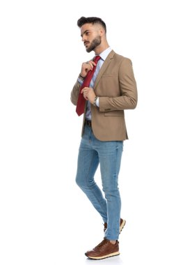 Kendine güveni olan sakallı bir adam kırmızı kravat takıyor, havalı davranıyor ve stüdyoda beyaz arka planda yürüyor.