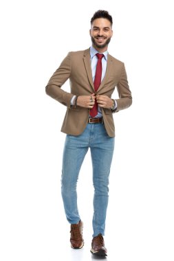 Kırmızı kravat düğmeli ceketli traşsız genç adam, gülümsüyor, mutlu ve stüdyoda beyaz arka planda yürüyor.