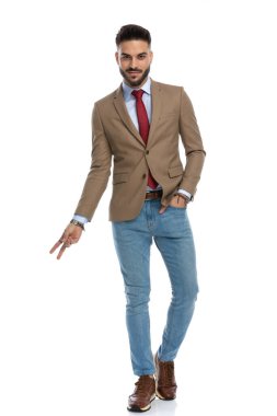 Kırmızı kravatlı yakışıklı genç adam gülümsüyor ve bir eliyle barış işareti yapıyor, diğerini cebinde tutuyor ve beyaz arka planda yürüyor.