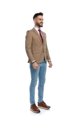 Kırmızı kravatlı ve ceketli gururlu sakallı adam beyaz arka planda poz verirken başka tarafa bakıyor ve gülümsüyor.