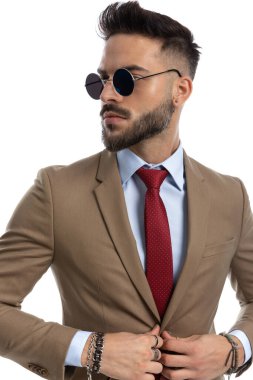 Ceketinin düğmelerini açarken ve stüdyoda beyaz arka planda poz verirken gözlüklü havalı sakallı adamın fotoğrafı.