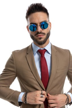 Retro güneş gözlüklü kendine güvenen bir adam ceket düğmelerinin üzerinde el ele tutuşuyor ve stüdyoda beyaz arka planda poz verirken ayarlıyor.