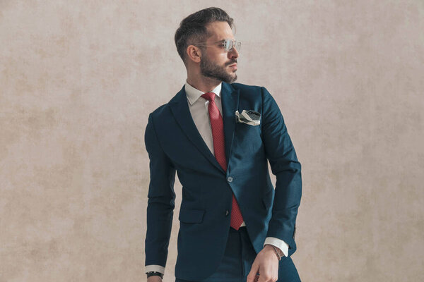 sexy bearded man wearing navy blue suit with red tie and handkerchief looking to side and posing on beige background in studio