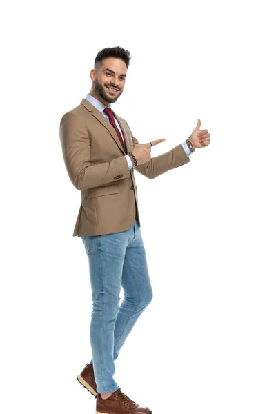 スタジオで白い背景を歩いている間に親指を立てジェスチャーと指を横に向けるジャケットの幸せな若い男の全身像 — ストック写真