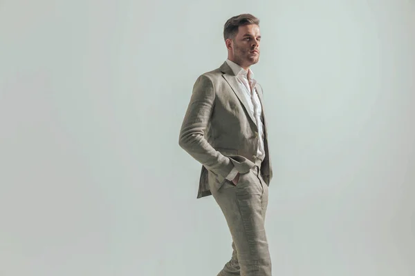 side view of confident young businessman with hands in pockets  looking to side, walking and posing in front of grey background in studio