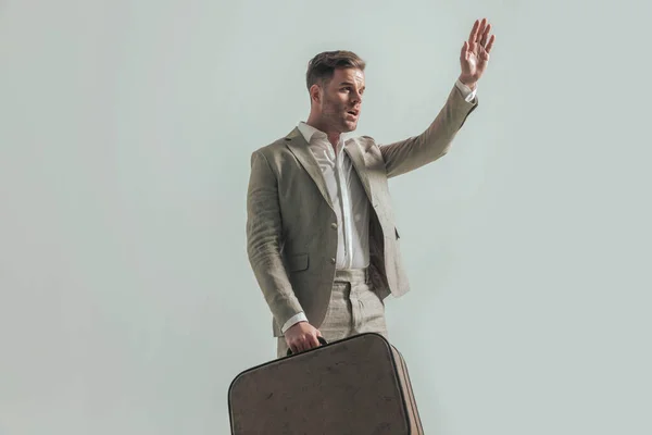 attractive guy with open collar shirt, holding luggage, looking to side and holding hand up getting a cab to airport, posing in front of beige background in studio