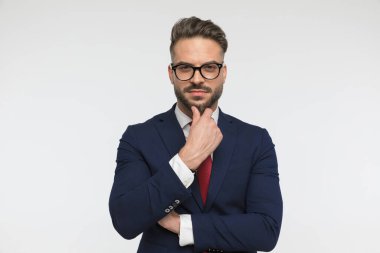 Gözlüklü, çenesine dokunan ve stüdyoda beyaz arkaplan önünde poz veren düşünceli bir adamın portresi.