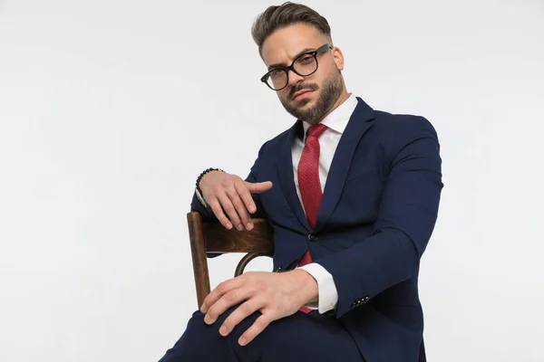 cool bearded businessman with glasses holding one arm over chair and touching knee while sitting in front of white backgrund in studio