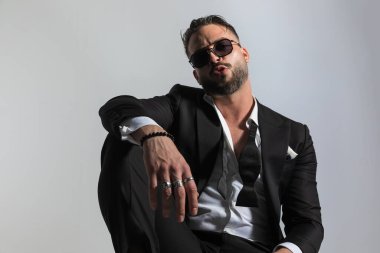 Siyah smokinli seksi adam moda pozunda kollarını tutuyor, öpücükler yolluyor ve stüdyodaki gri arka planda karşı konulmaz oluyor.
