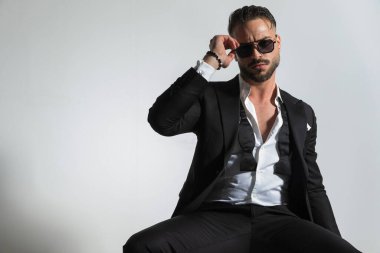 Siyah smokinli ve papyonlu şık bir iş adamı stüdyoda gri arka planda otururken ve poz verirken güneş gözlüğünü ayarlıyor.