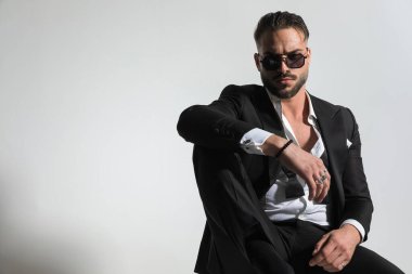 Smokinli, güneş gözlüklü, seksi ve zarif bir adam. Gri arka planda dirseğiyle poz veriyor.